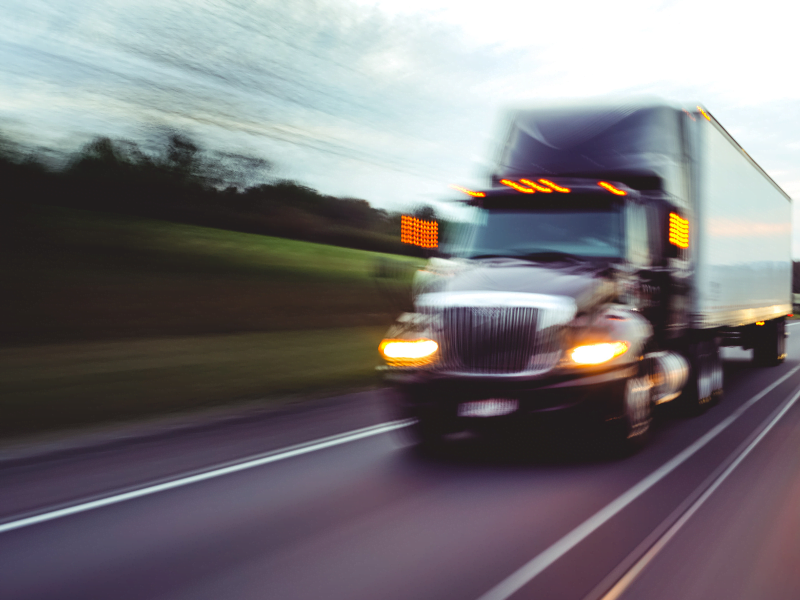 A semi-truck speeds down a highway, creating a motion blur effect, with green landscape in the background.