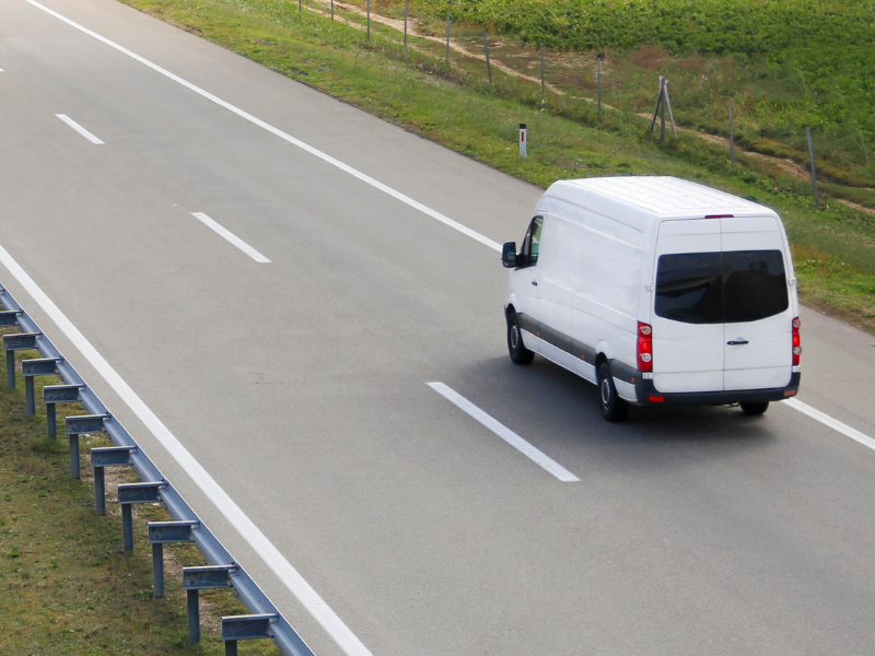 White van driving on an empty highway, surrounded by grassy areas and guard rails on a clear day.